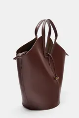 Bolso tipo shopper con forma de capazo, color marrón oscuro, confeccionado en material liso con pespuntes visibles en el cuerpo. Posee doble asa de mano y un asa bandolera ajustable. Incluye un bolso interior extraíble que se cierra mediante fruncido y un cierre principal con botón imantado.