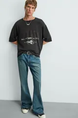 Camiseta de punto con corte boxy fit, cuello redondo y manga corta. Color negro con efecto lavado y terminaciones irregulares. Presenta un estampado frontal con texto y un gráfico circular en tonos blanco y rojo.