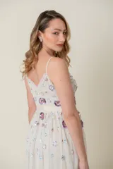Vestido midi de tul color marfil con escote en V y tirantes finos. El corpiño y la falda están adornados con lentejuelas y apliques bordados en tonos pastel como lila, rosa y azul, simulando flores. La falda es voluminosa y con capas de tul.