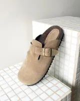 Zueco de cuero gamuzado color beige, con diseño cerrado en la punta y ajuste mediante una correa con hebilla metálica en el empeine. Presenta una plantilla anatómica de corcho y suela dentada negra.