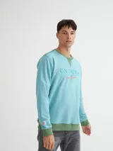 Buzo azul con cuello redondo, mangas largas y puños y bajo acanalados. Presenta bloque de color naranja en hombros y laterales, y estampado frontal con texto en color borgoña.