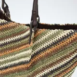 Bolso tipo capazo grande de tejido trenzado con diseño de rayas diagonales multicolor en tonos verdes, marrones, blancos y negros. Cuenta con doble asa de hombro en cuero sintético marrón.
