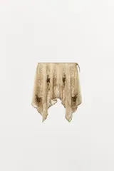 Falda corta de tiro bajo confeccionada en tul color beige, con capas, bajo asimétrico, bordados florales oscuros con lentejuelas en contraste y cierre lateral con nudo.