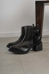 Bota corta de cuero negro con acabado combinado mate y charol. Presenta una tira decorativa con tachas metálicas y hebilla en el tobillo, tacón cuadrado robusto y suela con plataforma.