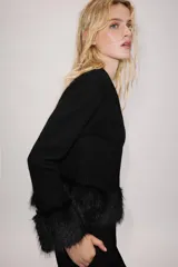 Chaqueta de punto negra con cuello redondo y manga larga, con detalles de pelo sintético a tono en los puños y en el bajo. Cuenta con cierre frontal con botones a presión ocultos.
