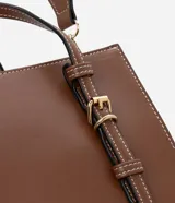 Cartera satchel mediana de color marrón, confeccionada en poliuretano con diseño estructurado, costuras en contraste y herrajes metálicos dorados. Cuenta con doble asa de mano, correas decorativas verticales con hebillas en la parte frontal y una correa larga ajustable y desmontable para llevar al hombro o cruzada.