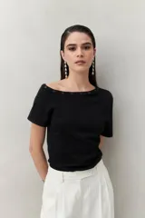 Top de tejido negro ajustado al cuerpo, con escote barco adornado con botones metálicos oscuros. Tiene mangas cortas.