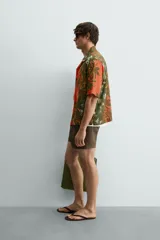 Camisa de corte relajado confeccionada en tejido fluido con mezcla de viscosa y lino. Presenta cuello bowling, manga corta y cierre frontal con botones. Diseño con estampado floral en tonos verdes y naranjas.
