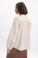 Camisa de manga larga color amarillo pálido con estampa boho a rayas verticales compuestas por pequeños motivos florales. Posee cuello alto con volados y cordones para atar en el frente, y mangas abullonadas con puños ajustados.