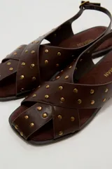 Sandalias planas de cuero sintético marrón, con tiras cruzadas en el empeine y tachas metálicas doradas distribuidas por toda la capellada. Cuenta con tira ajustable en el talón con hebilla.