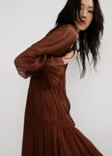 Vestido midi marrón de viscosa transparente, con diseño plisado, cuello alto fruncido y mangas largas abullonadas con puños elásticos. Presenta detalles de costuras fruncidas en la cintura y falda escalonada con vuelo.