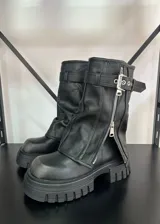Botas de caña media color negro, con suela track dentada, cierre frontal con cremallera metálica y tira con hebilla ajustable en la parte superior.