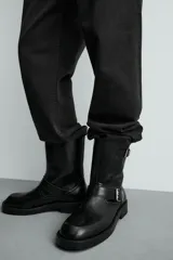 Bota de media caña en color negro, con diseño de horma cuadrada y suela con ligero tacón. Presenta dos cintas ajustables con hebillas metálicas, una en la parte superior de la caña y otra en la zona del empeine.