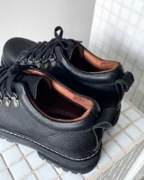 Zapatos tipo borceguíes bajos de cuero vacuno negro, con cordones y ganchos metálicos. Presentan suela track dentada y costuras visibles en el borde de la suela.