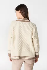 Sweater de punto estilo preppy en color crudo con detalles en marrón. Presenta cuello en V con ribete a rayas, tejido con textura de ochos y cordones laterales decorativos. Posee puños y ruedo acanalados con diseño a rayas.