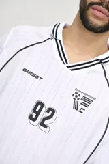 Camiseta de fútbol blanca con cuello en V y mangas largas con puños a rayas negras. Tiene un logo de la marca en el pecho y el número 92 en el centro.