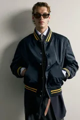 Cazadora bomber estilo universitario, de corte relaxed fit y tejido técnico azul marino, con cuello, puños y dobladillo de rib elástico a rayas amarillas. Cuenta con bolsillos delanteros de plastrón y cierre frontal con botones a presión.