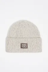 Gorro tipo beanie de punto texturizado en color gris claro con destellos plateados. Presenta un parche rectangular en la parte frontal con el logo de la marca en color marrón.