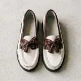 Mocasín de estilo náutico confeccionado en cuero, combinando una base de cuero con textura de pelo en color negro y detalles de cuero con textura croco en color marrón. Presenta cordones decorativos con borlas laterales y suela plana de goma.