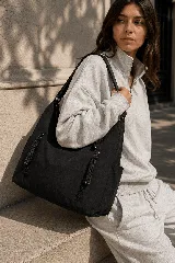 Cartera tipo hobo color negro, confeccionada en tela suave y espaciosa, con dos bolsillos frontales verticales con tiradores trenzados y correa ajustable para el hombro.