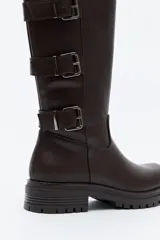 Bota de caña alta tipo biker en símil cuero color marrón oscuro. Presenta múltiples tiras horizontales con hebillas metálicas laterales y una suela robusta con relieve.