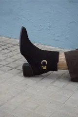 Bota corta de cuero color marrón, con taco cuadrado de madera y punta fina. Presenta una tira decorativa con hebilla metálica dorada alrededor del tobillo.