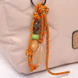Bandolera tipo shopper color beige con diseño acolchado y pespuntes geométricos. Tiene doble asa de mano en tono tostado y una correa larga regulable y extraíble. Incluye cierre superior, bolsillo frontal con cierre adornado con un llavero de cordón naranja con cuentas y un parche decorativo.