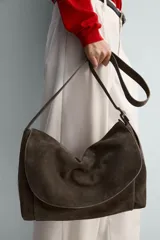 Bolso tipo bandolera de piel con acabado serraje, color marrón oscuro. Presenta un compartimento principal con cierre de solapa y cremallera, y un bolsillo interior con cierre de cremallera. Incluye un asa de hombro ajustable de piel.