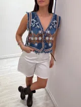 Chaleco corto en denim azul con detalle desflecado y bordado artesanal en tonos celeste y dorado. Estilo boho chic con escote en V, ideal para combinar con prendas básicas y elevar tu look.