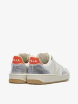 Championes Veja modelo V-10, con diseño de paneles en cuero blanco y cuero metalizado plateado. Presentan el logo lateral en color crema, detalle de talón en color rojo con la marca estampada y suela de caucho color caramelo.