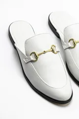 Mules chatitas de cuero vacuno color blanco, con punta redondeada y detalle de hebilla metálica dorada.