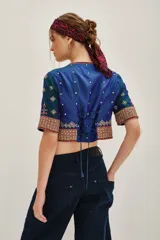 Camisa de seda con diseño tipo chaqueta corta, de color azul petróleo con bordados decorativos en tonos dorados y rojos. Presenta mangas largas abullonadas de color crema y cierre frontal.
