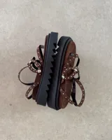 Sandalia de plataforma negra con tiras decoradas con tachas metálicas plateadas. Posee un diseño tipo ojota con ajuste de pulsera al tobillo mediante hebilla.
