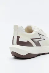 Zapatilla deportiva de diseño dinámico, confeccionada en material técnico con paneles en contraste. Presenta un diseño lateral con forma de rayo en tono marrón, ajuste con cordones y suela robusta con relieve.