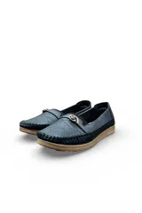 Mocasines negros de cuero sintético con perforaciones en la capellada, costuras visibles en relieve y una tira decorativa en el empeine con una hebilla metálica dorada. Tienen suela plana de goma marrón clara.