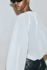 Blusa de crepé de gasa color blanco, con cuello mao, abertura frontal con tiras para anudar y detalle de volados verticales. Presenta mangas largas tipo globo de corte raglán con puños elásticos y terminación en volados.