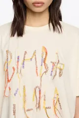 Camiseta de manga corta, corte oversize, color marrón oscuro. Presenta un estampado frontal con el texto "BIMBA Y LOLA" realizado con hilos de colores vibrantes que cuelgan, simulando un bordado texturizado.