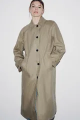 Trench coat de corte largo y diseño minimalista, confeccionado en tejido encerado color beige. Presenta cuello solapa, cierre frontal con botones oscuros y bolsillos laterales integrados.