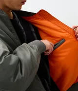 Campera tipo bomber de color verde militar, con cierre frontal, cuello, puños y cintura en tejido rib. Presenta bolsillos frontales con solapa y botón a presión, además de un bolsillo utilitario en la manga.
