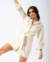 Vestido camisero corto de denim azul oscuro, con cuello clásico, mangas 3/4 con puños abotonados, cierre frontal con botones color cobre, cinturón de lazo y bolsillos con solapa en el pecho.