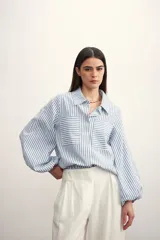 Camisa celeste a rayas blancas, con cuello clásico, cierre frontal con botones, mangas largas abullonadas con puños elásticos y dos bolsillos de parche en el pecho.