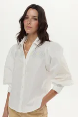Camisa blanca de algodón con mangas abullonadas, cuello camisero y bordados geométricos y florales en tonos celestes y amarillos en el frente y las mangas.