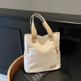 Cartera tipo tote de lona color marrón con asas de mano en color beige. Cuenta con un bolsillo frontal, bolsillos laterales y un detalle de tirador de cierre en cordón blanco.