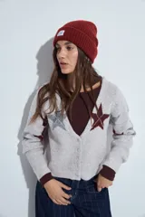 Cardigan corto tejido en color gris, con escote en V y cierre frontal de botones. Presenta un diseño estampado con estrellas y líneas geométricas en tonos oscuros y rojizos, con mangas largas y puños acanalados.