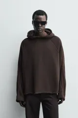 Sudadera de corte boxy fit en color marrón oscuro, con capucha, mangas largas y bandas laterales a contraste. Presenta un diseño de bajo con acabado irregular deshilachado.