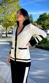 Cardigan de punto en color blanco con detalles en negro en los bordes, cuello en V, cierre frontal con botones dorados y bolsillos delanteros.