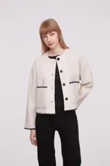 Chaqueta corta de paño color marfil, con cuello redondo, cierre frontal de botones negros y dos bolsillos frontales. Presenta detalles de costuras decorativas en contraste negro en los bordes de los bolsillos, puños y ruedo.