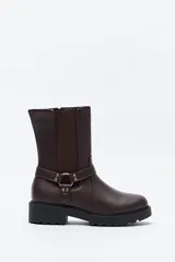 Bota tipo biker de caña media en cuero sintético negro, con panel elástico lateral, suela gruesa dentada y arnés decorativo con herraje metálico.