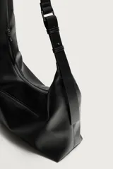 Bolso de hombro asimétrico color negro con acabado liso. Cuenta con un compartimento principal con cierre de cremallera y un bolsillo frontal con cremallera. Incluye asa de hombro ajustable.