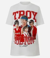 Remera de manga corta color blanco roto con cuello redondo. Presenta un estampado grande en el frente con la palabra "TROY" en letras rojas con textura de puntos blancos, y un collage de cuatro imágenes del actor Zac Efron en su papel de Troy Bolton de High School Musical, rodeado de estrellas rojas.